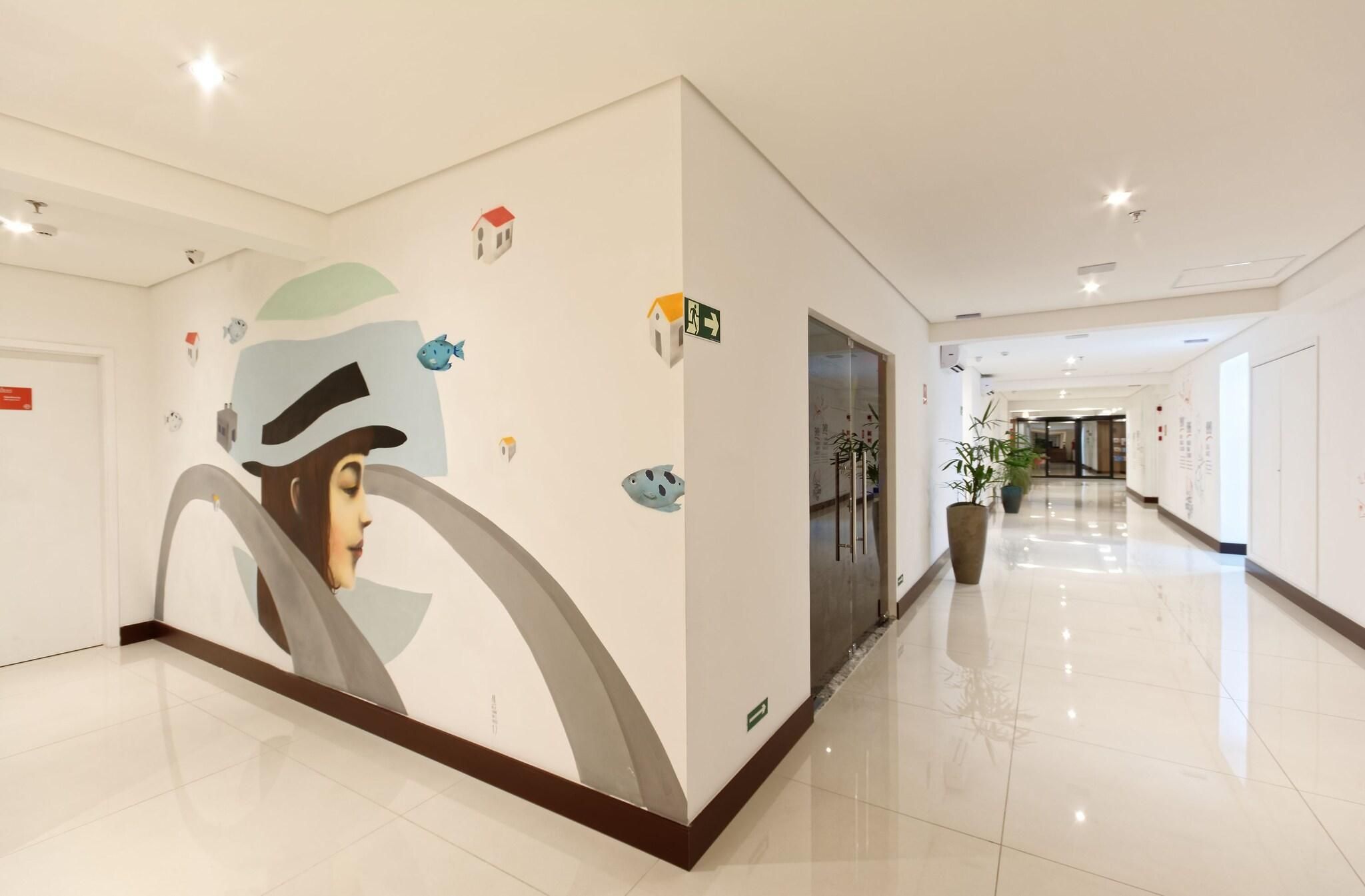 Corredor de hotel com um grande mural na parede, um vaso de planta e uma porta com sinalização de saída de emergência.