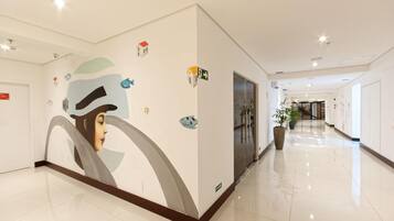 Corredor de hotel com um grande mural na parede, um vaso de planta e uma porta com sinalização de saída de emergência.