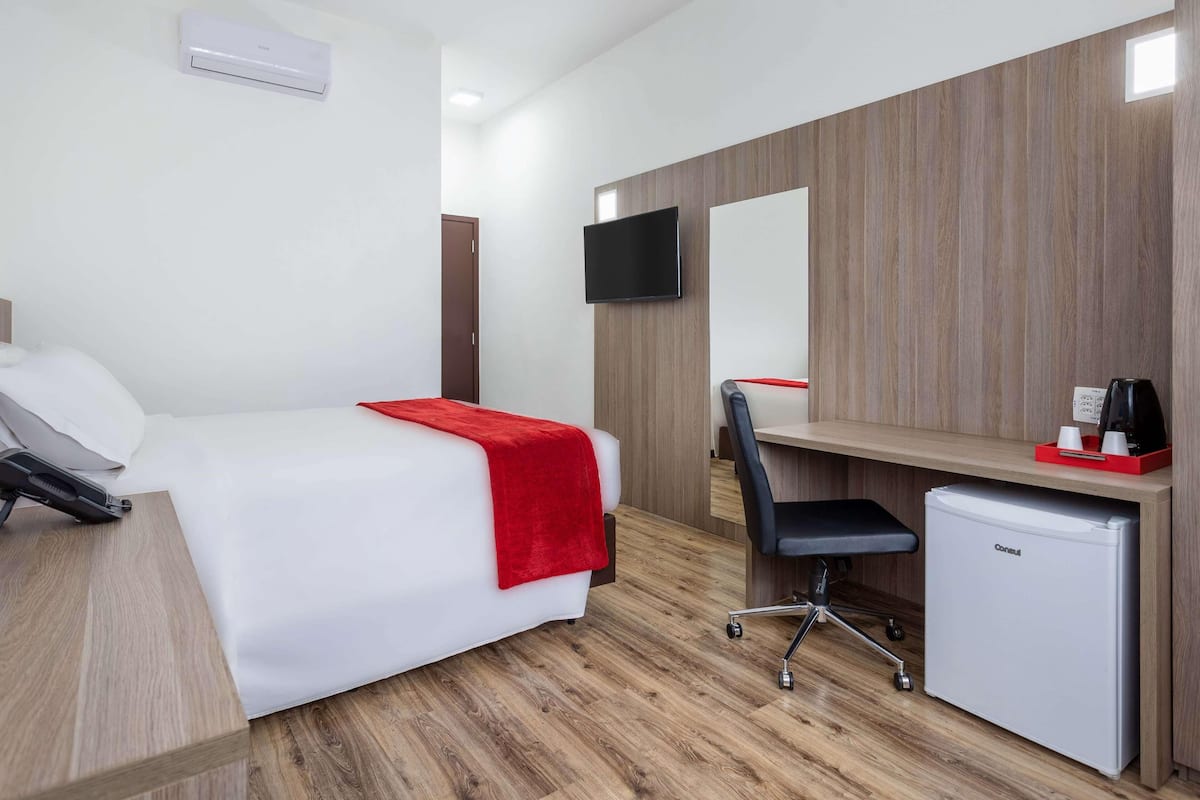 Quarto de hotel com cama, escrivaninha, cadeira, mini-geladeira, televisão e ar-condicionado.