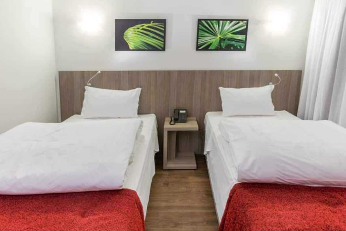 Quarto de hotel com duas camas de solteiro, cabeceira de madeira, mesa de cabeceira com telefone e duas imagens penduradas na parede.