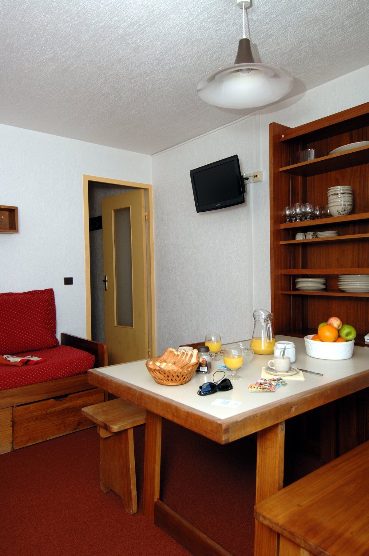 A compact room with a wooden table, a television mounted on the wall, a red sofa, and a breakfast setup with fruits and beverages.