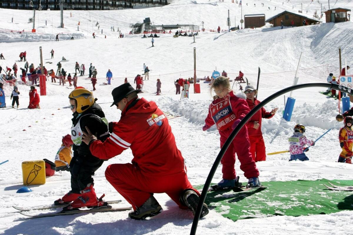A snowy ski slope with people skiing and standing, buildings in the background, and a clear sky.