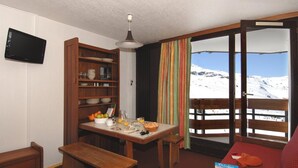 A room with a wooden table set for breakfast, a television mounted on the wall, and a view of a snowy mountain through a window. - Residence Odalys Tourotel (Les Belleville)