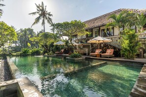 Une piscine entourée de chaises longues et de parasols, bordée d’une végétation tropicale et d’un bâtiment doté d’un toit de chaume.
