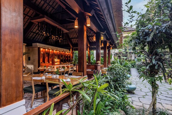 Un restaurante al aire libre con mesas y sillas de madera, un área de estar cubierta y abundante vegetación.