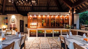 A restaurant with a high ceiling, patterned floor tiles, and a bar area with shelves of bottles and glasses. - Radha Phala Resort and Spa (Ubud)