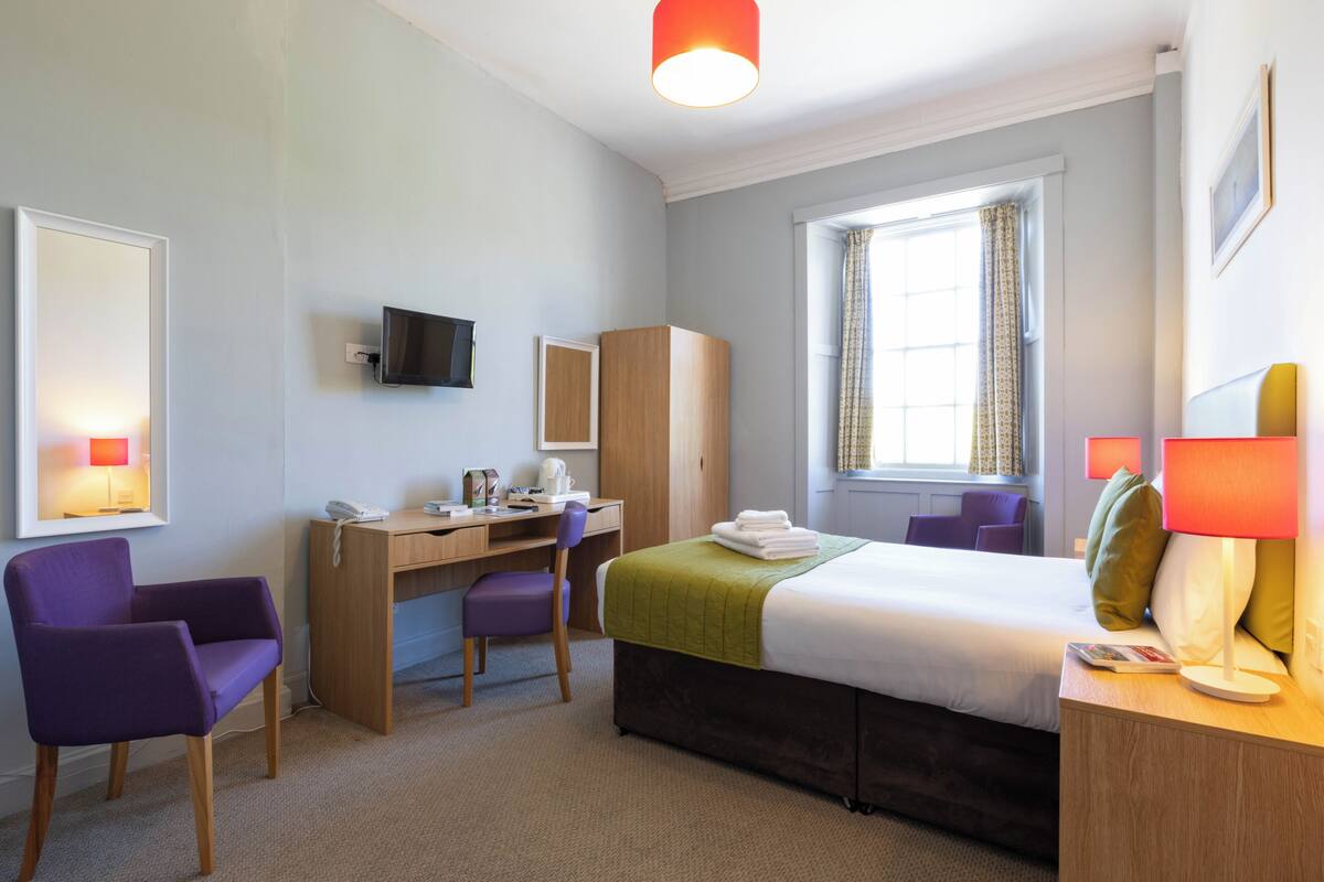 A hotel room with a bed, desk, chair, television, and a window with curtains.