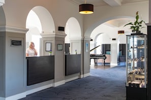 Eine Hotelhalle mit Empfangstresen, einem Klavier und einer Vitrine mit Ausstellungsstücken.