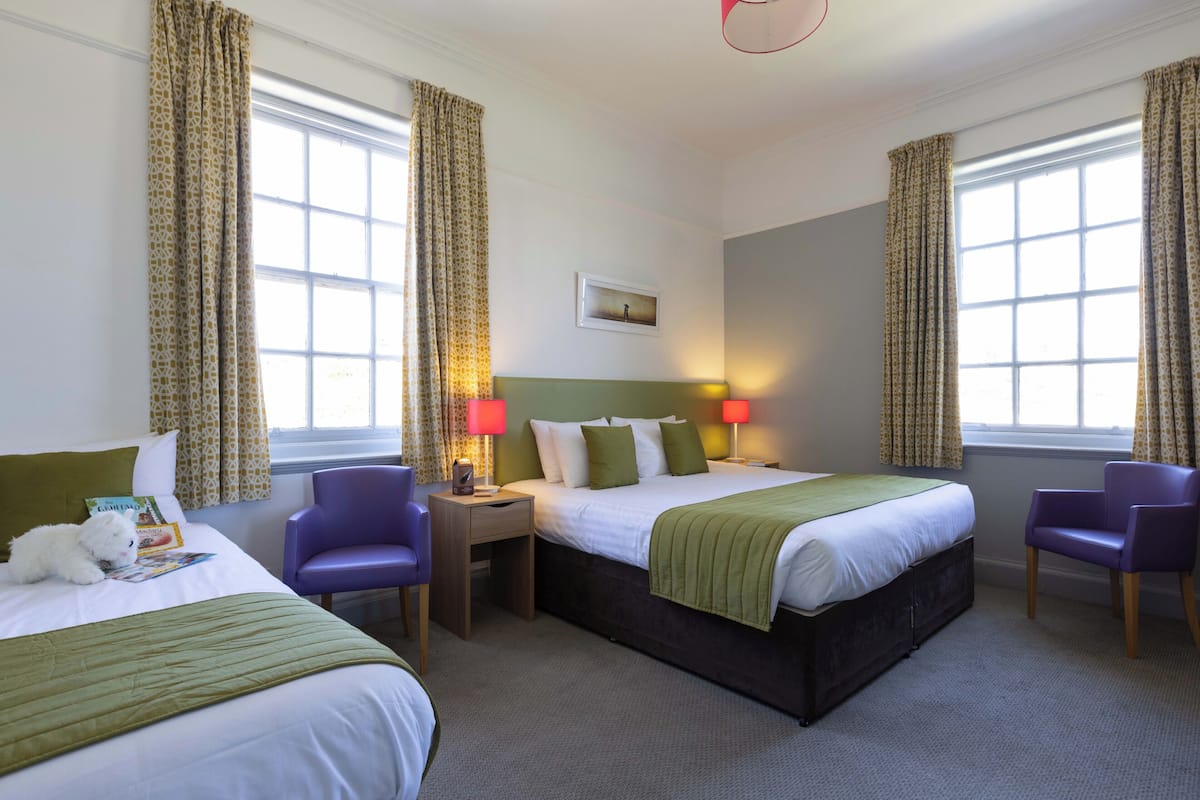 A hotel room with two beds, a desk, two chairs, a lamp, and a window with curtains.