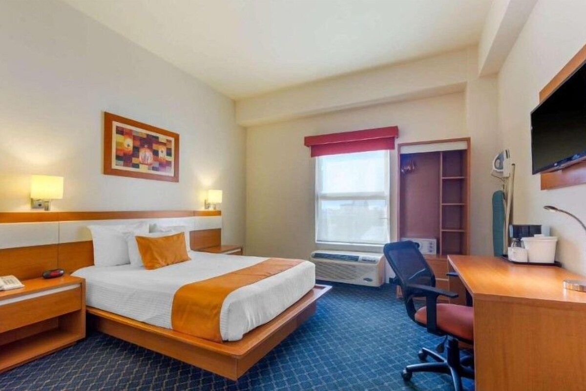 A hotel room with a bed, bedside lamps, a desk, a chair, a TV, and a window with a view.