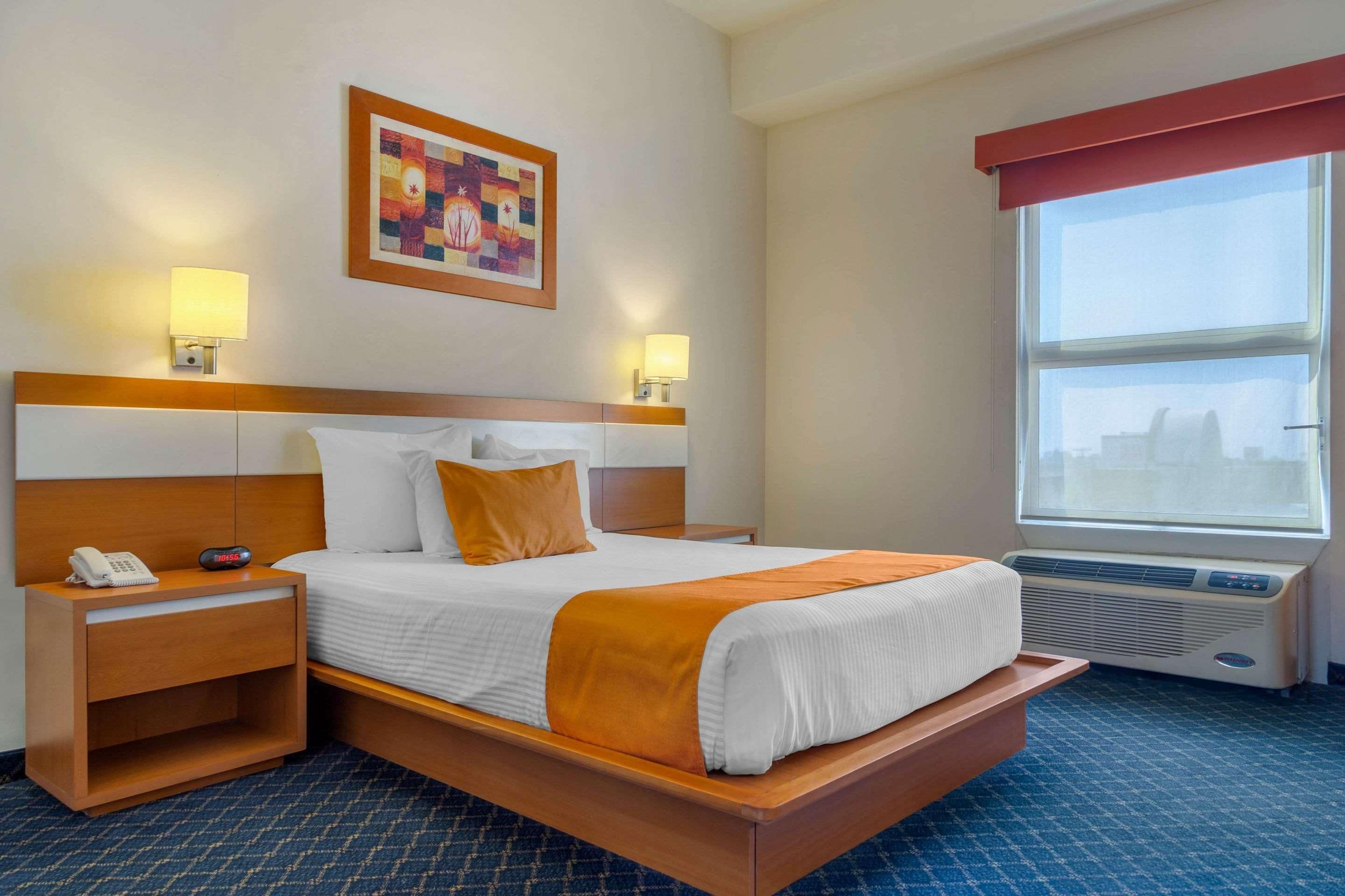 A hotel room with a bed, bedside lamps, a telephone, a painting, and a window with blinds.