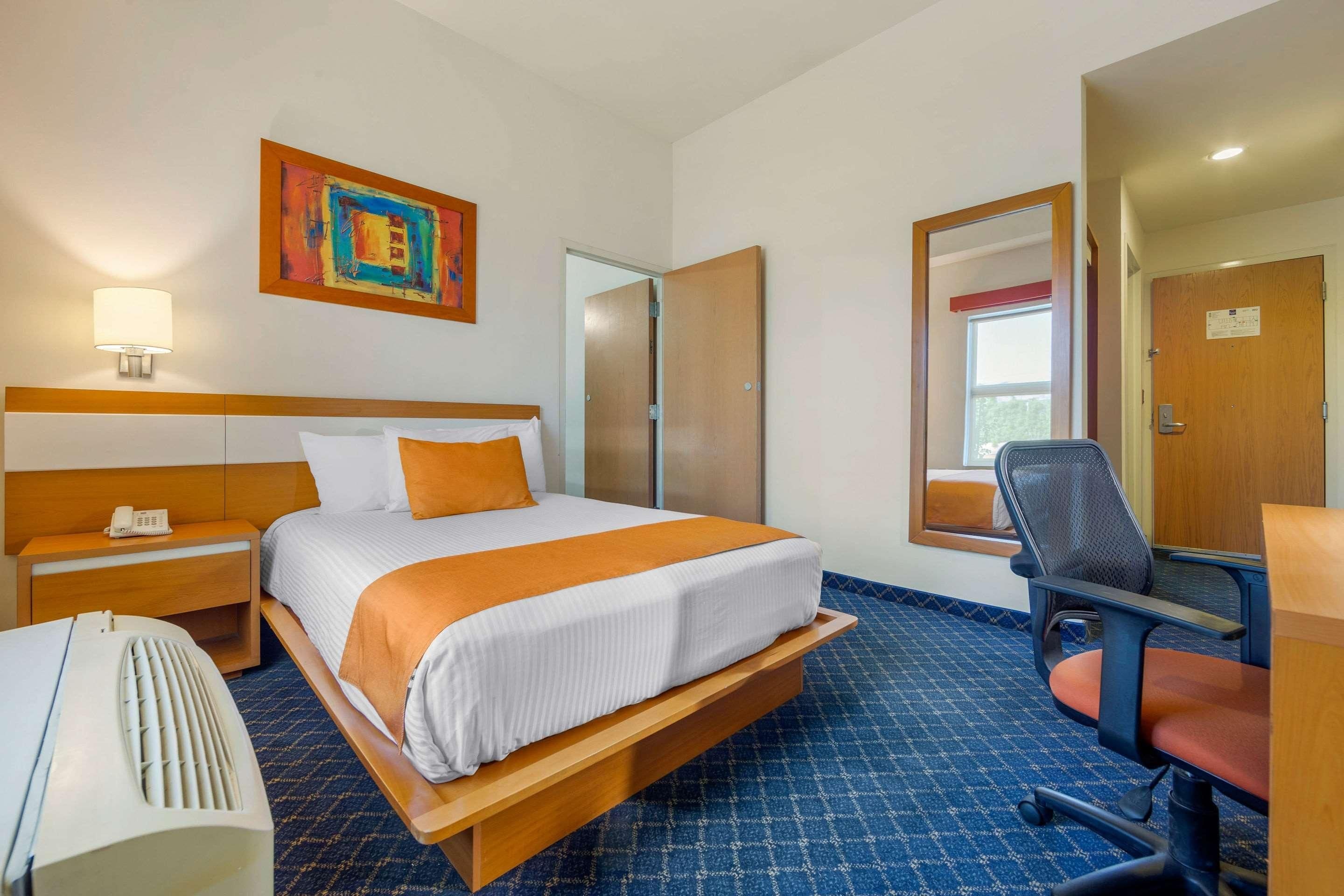 Una habitación de hotel con cama, mesita de noche, lámpara, silla y un cuadro en la pared.