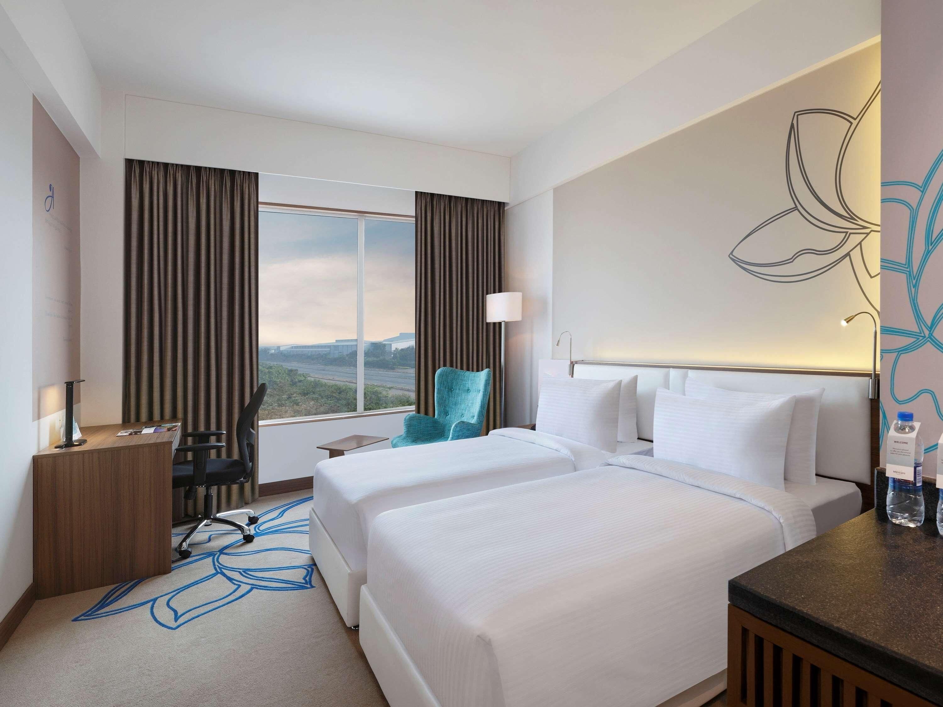 Quarto de hotel com duas camas, uma escrivaninha, uma cadeira, uma janela grande com cortinas e uma parede com um desenho floral.
