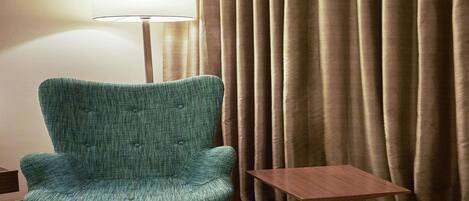 A teal armchair with wooden legs, a small wooden table, and a floor lamp in a room with beige curtains.