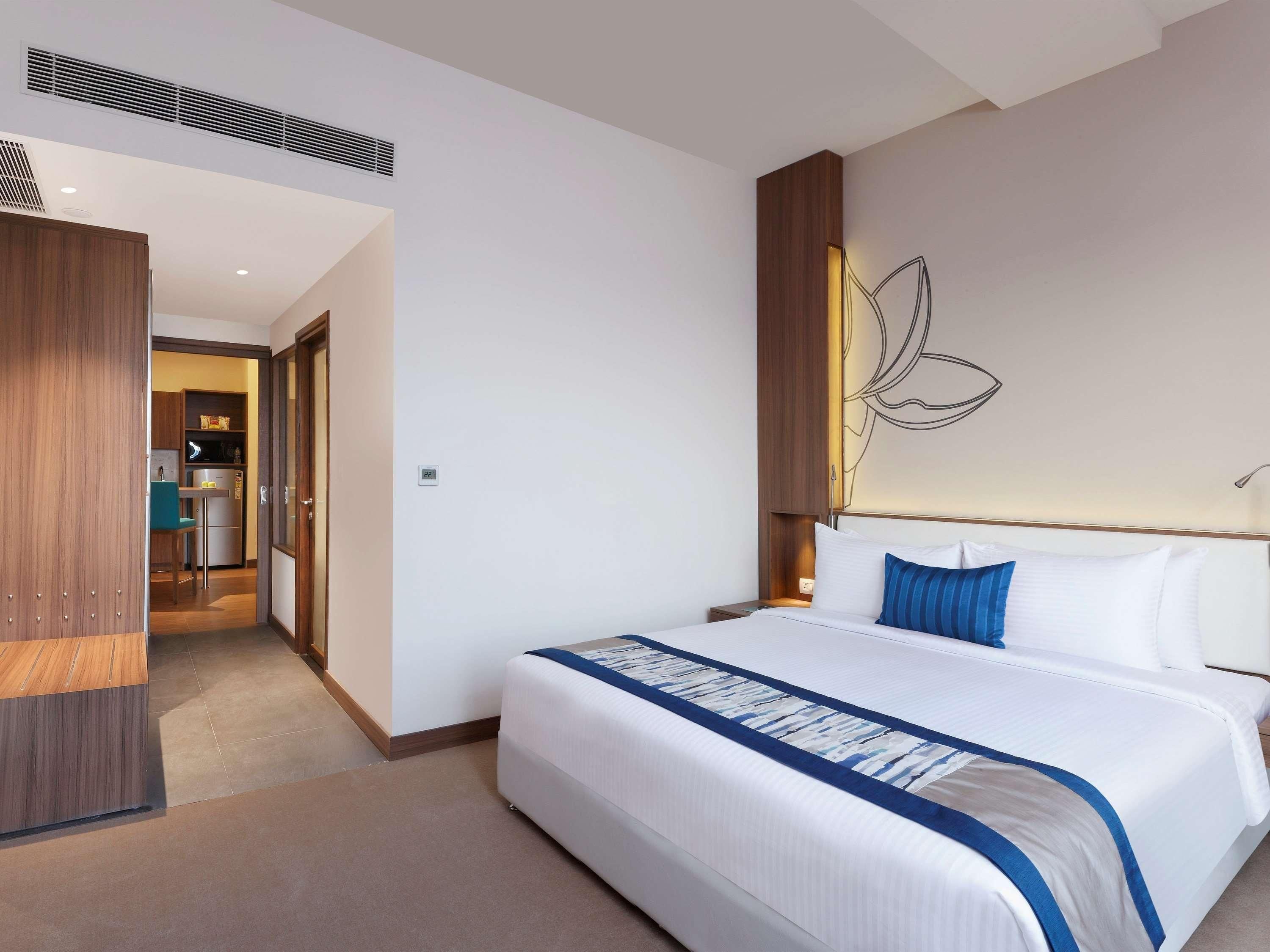 A hotel room with a bed, a wooden wardrobe, a kitchenette, and a wall with a light fixture.