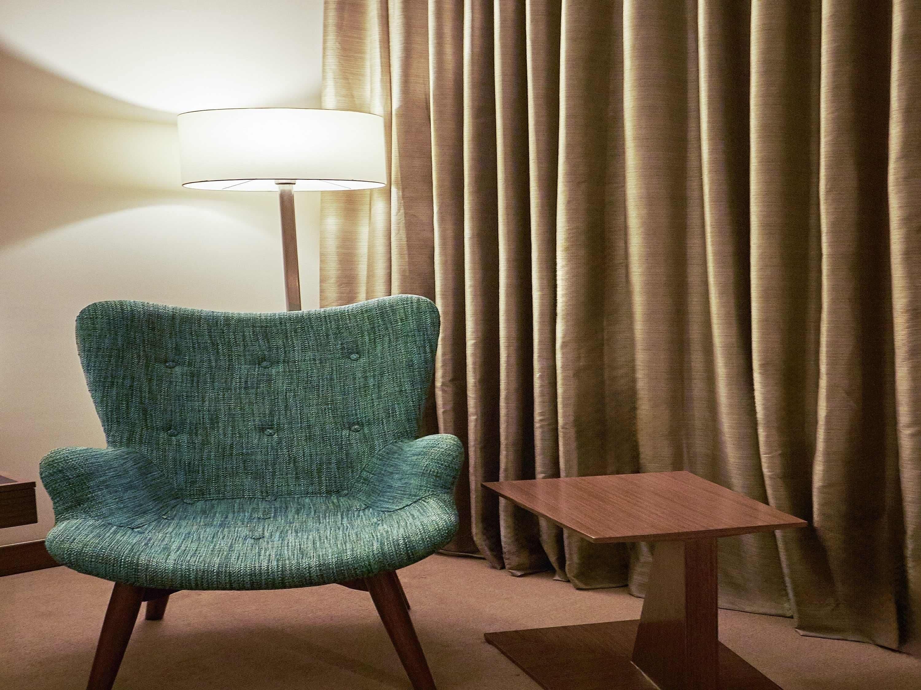 A teal armchair, a small wooden table, and a floor lamp in a room with a curtain backdrop.