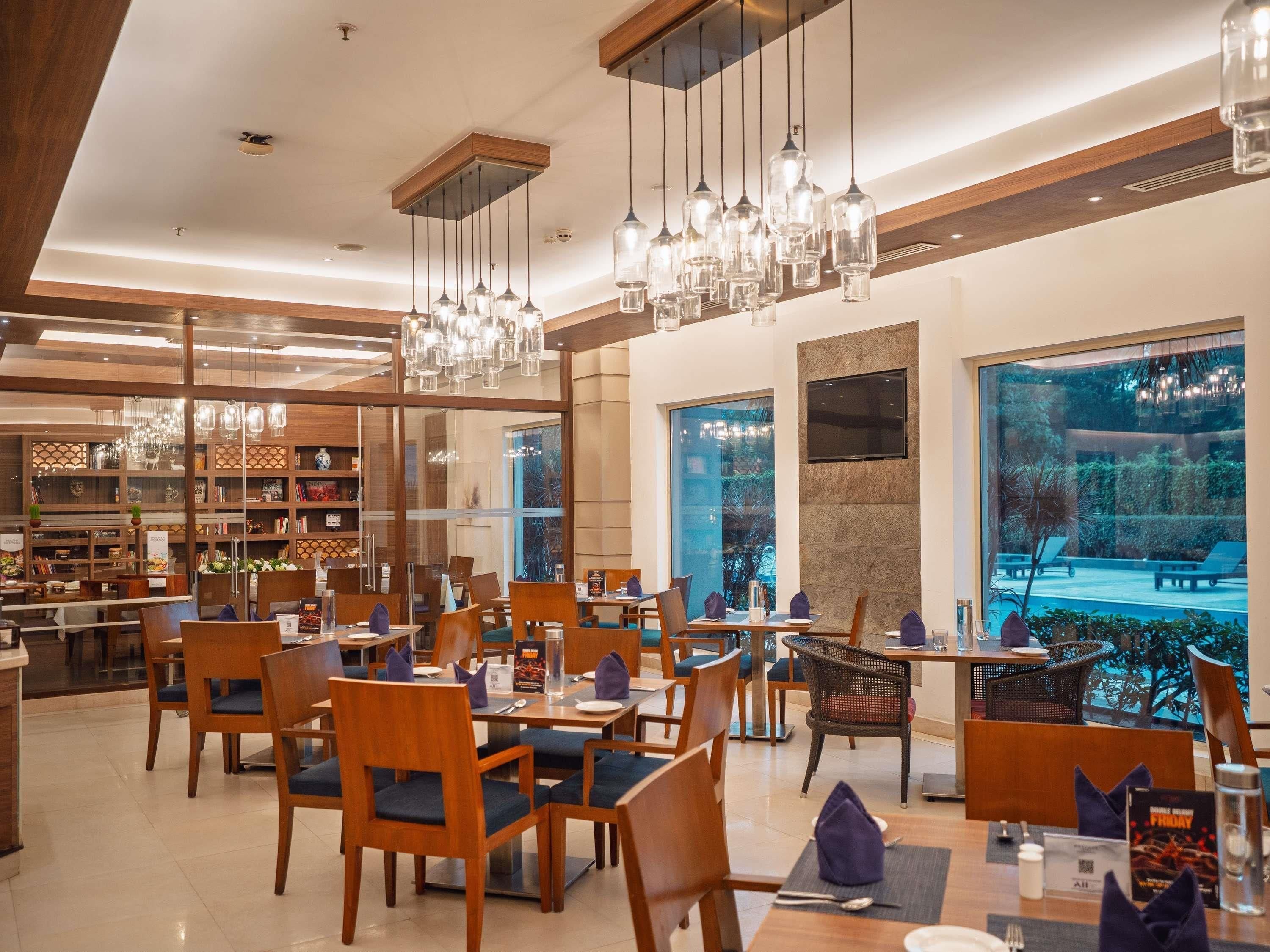 A modern dining area with multiple tables and chairs, a large window with a view of greenery, and hanging pendant lights.
