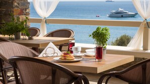 Una mesa de comedor con un plato de comida, una taza y un vaso de bebida roja, frente a una vista del mar y un yate.
