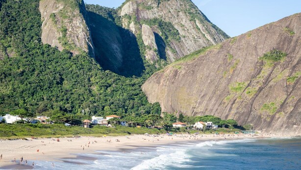 Ein Küstenstrand mit einem bergigen Hintergrund, Häuser entlang der Uferlinie und Menschen, die den Strand genießen.