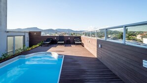 Ein Pool auf dem Dach mit Holzterrasse und Liegestühlen, umgeben von einer Holzverkleidung und einem Glasgeländer.