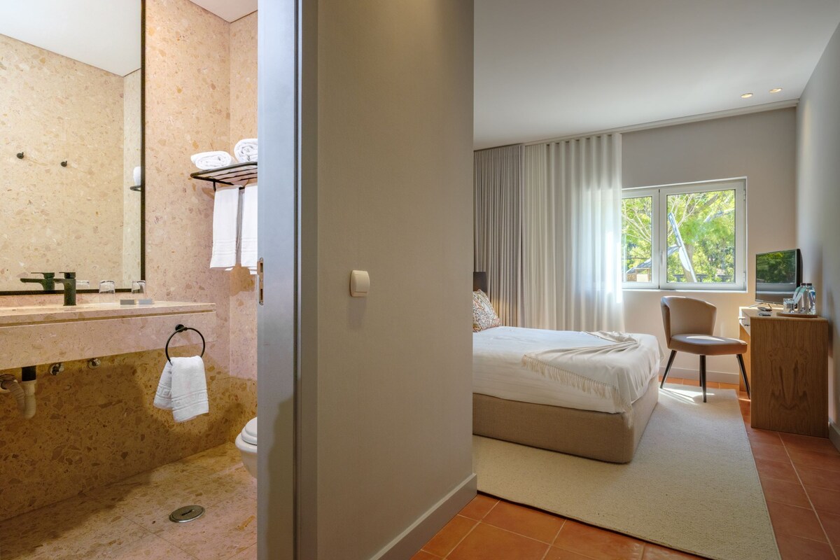 Quarto de hotel com banheiro, cama, escrivaninha e janela com vista.