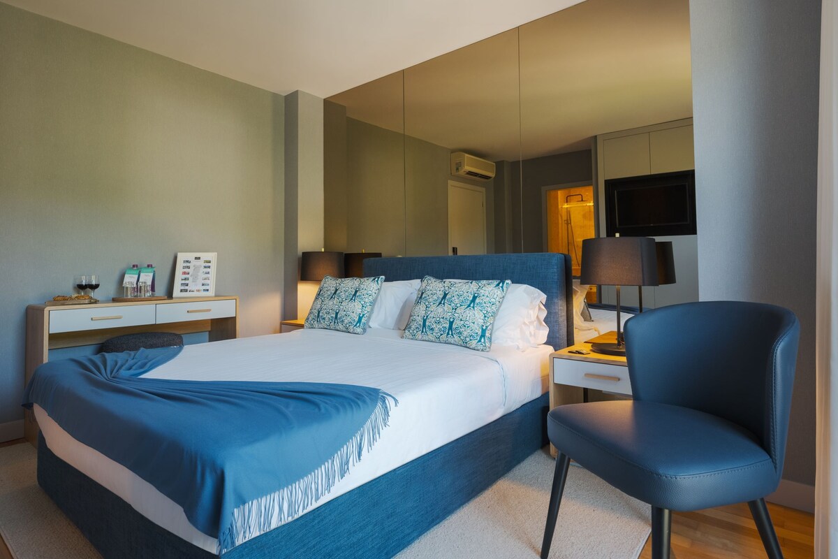 Una habitación de hotel con cama, mesitas de noche, una silla, televisión y espejo.