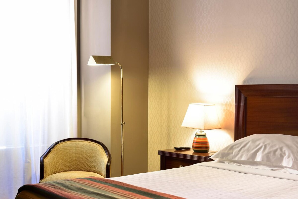 Habitación de hotel con cama, una silla, una mesita de noche con lámpara y ventana con cortinas.