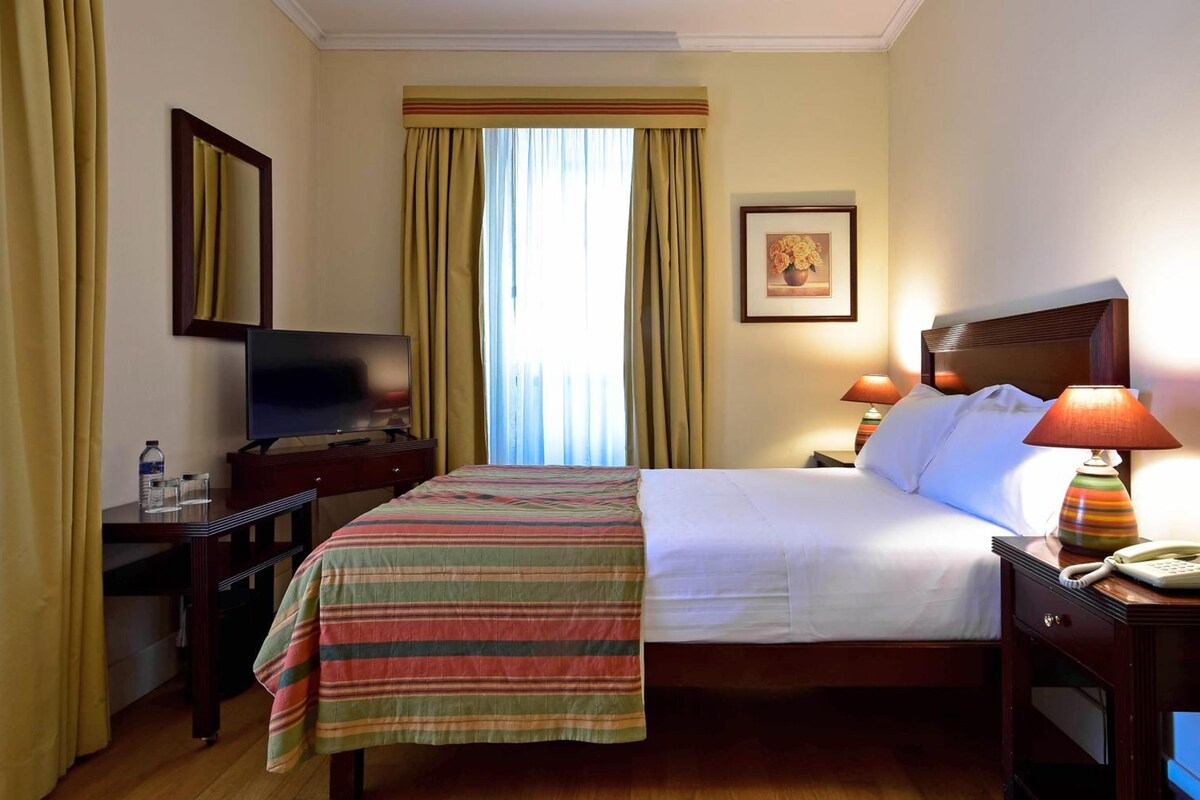Quarto de hotel com cama, escrivaninha com televisão, telefone e quadro na parede.
