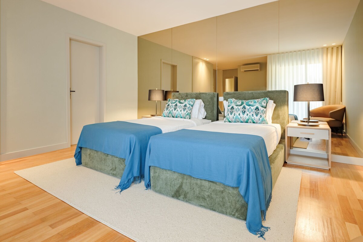 Uma cama de casal com cabeceiras combinando, uma mesa de cabeceira com abajur, um espelho e uma porta.
