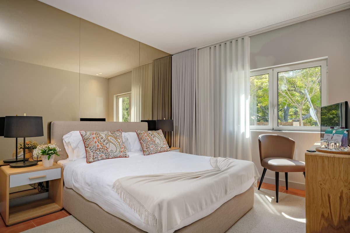 Una habitación de hotel moderna con una cama grande, lámparas de noche, un escritorio con una silla y una ventana con vista a árboles.