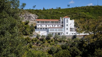A modern building with multiple floors, surrounded by greenery and a rocky hill.