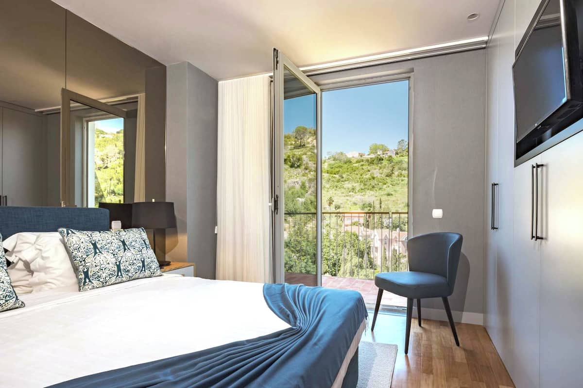 Un dormitorio moderno con una cama grande, una silla, un televisor y un balcón con vistas a las colinas.