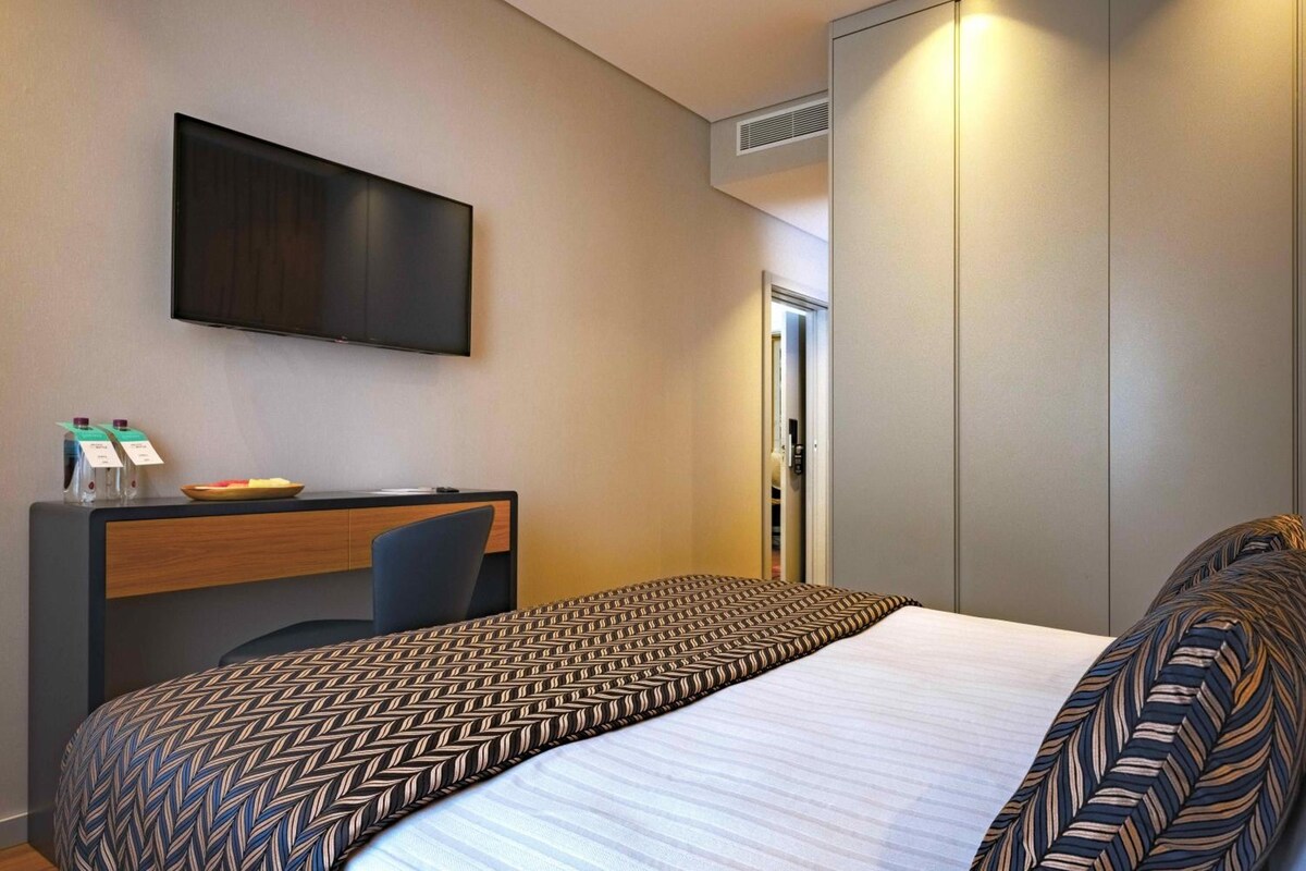 Una habitación de hotel con cama, escritorio, silla, televisor y armario.