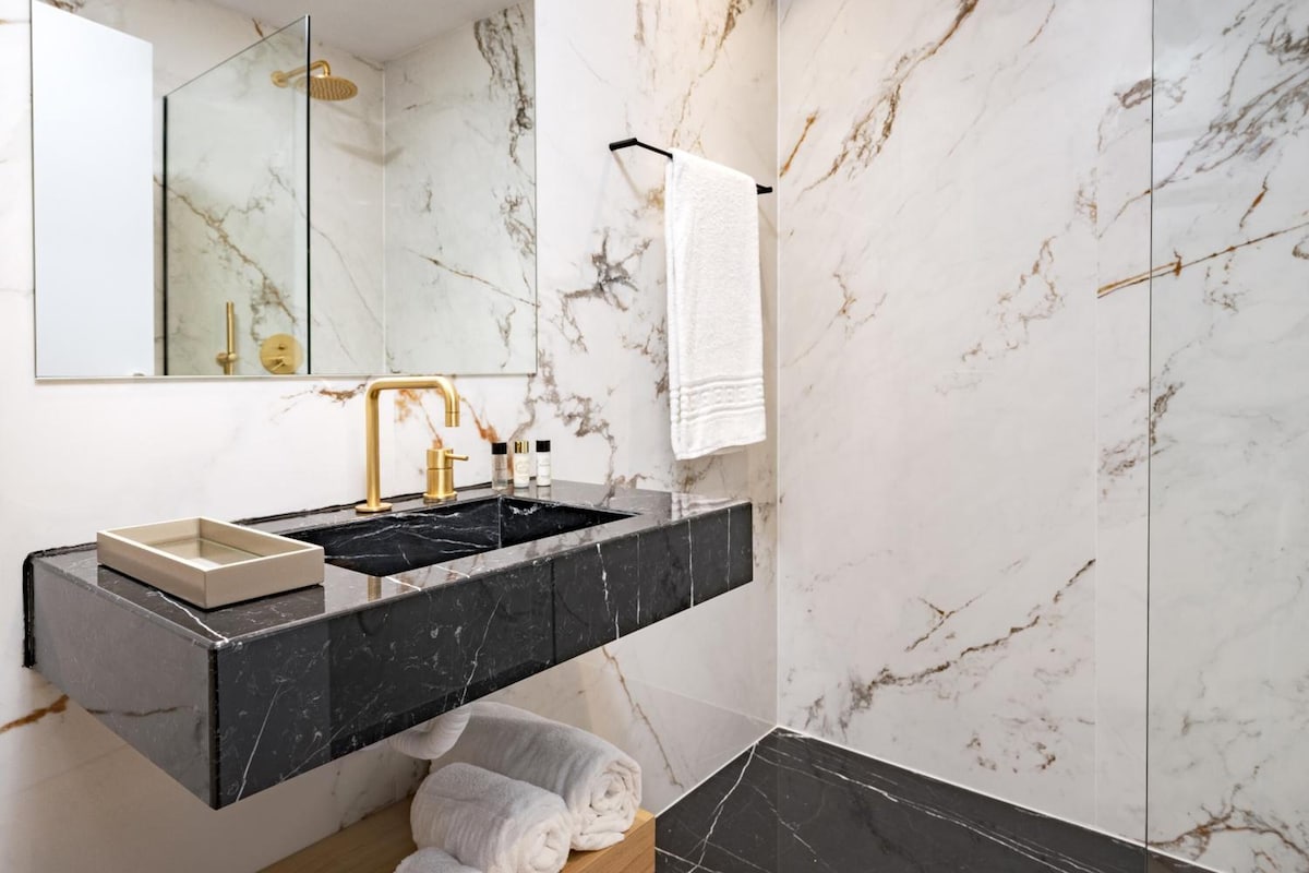 Un baño moderno con pared de mármol, un lavamanos negro con detalles dorados y una ducha con mampara de vidrio.