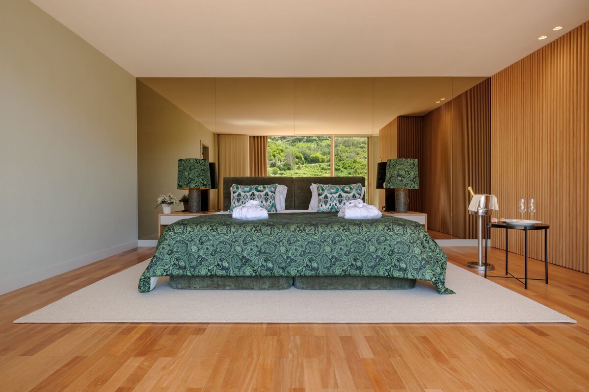 Quarto moderno com uma cama grande, duas mesas de cabeceira e vista para o verde lá fora.
