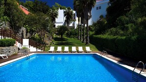 Una piscina con sillas de descanso blancas, rodeada de exuberante vegetación y un edificio blanco al fondo.
