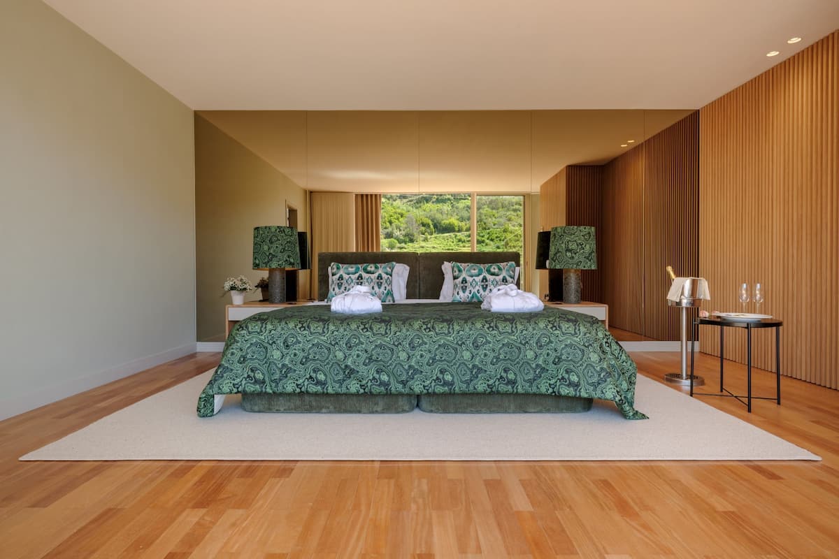 Un dormitorio moderno con una cama grande, dos mesitas de noche y una mesita auxiliar, con vistas a la vegetación exterior.