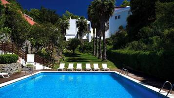 A swimming pool with white lounge chairs, surrounded by lush greenery and a white building in the background.