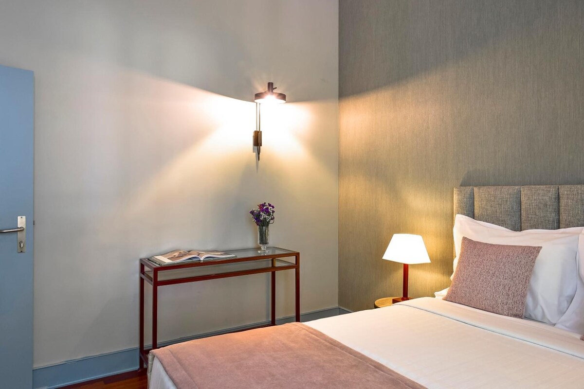 Habitación de hotel con cama, mesita de noche, lámpara y aplique de pared.