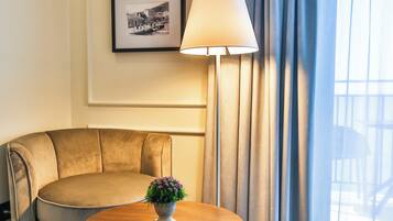 A hotel room with a round wooden table, a beige sofa, a floor lamp, and two framed pictures on the wall.