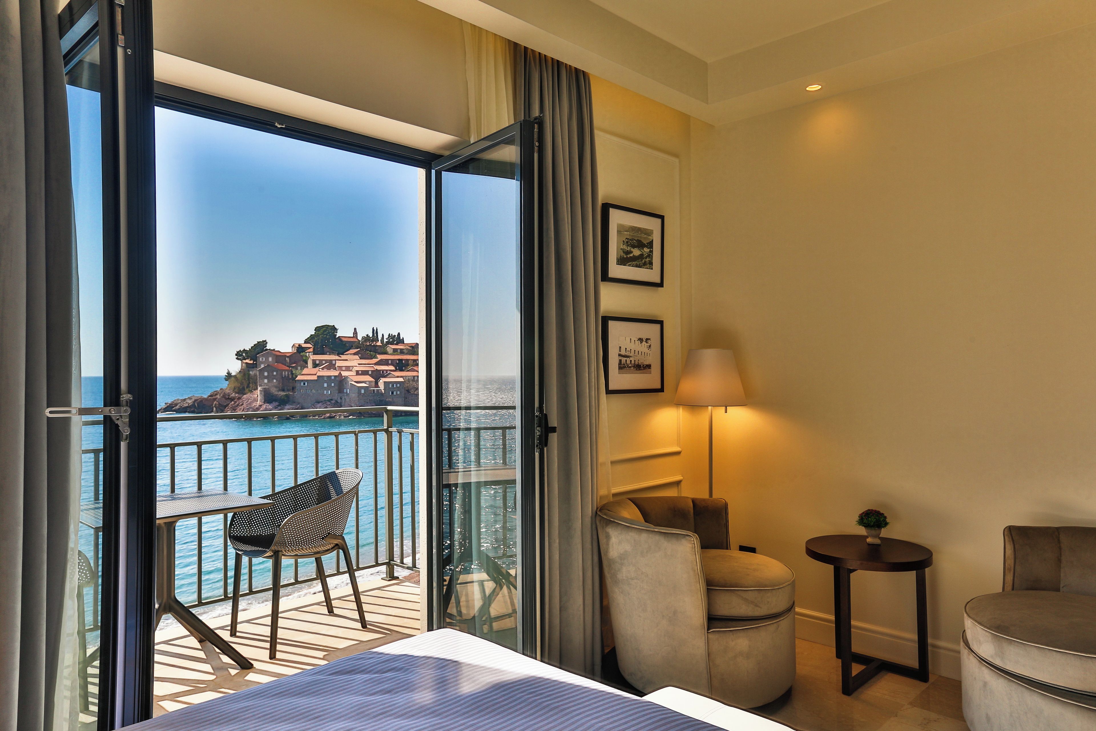 A hotel room with a balcony, a bed, a chair, a table, and a lamp.