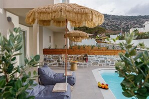 A poolside area with a stone wall, lounge chairs, and a thatched umbrella. - Anassa Stalis Suites Adults Only 16+ (Hersonissos)