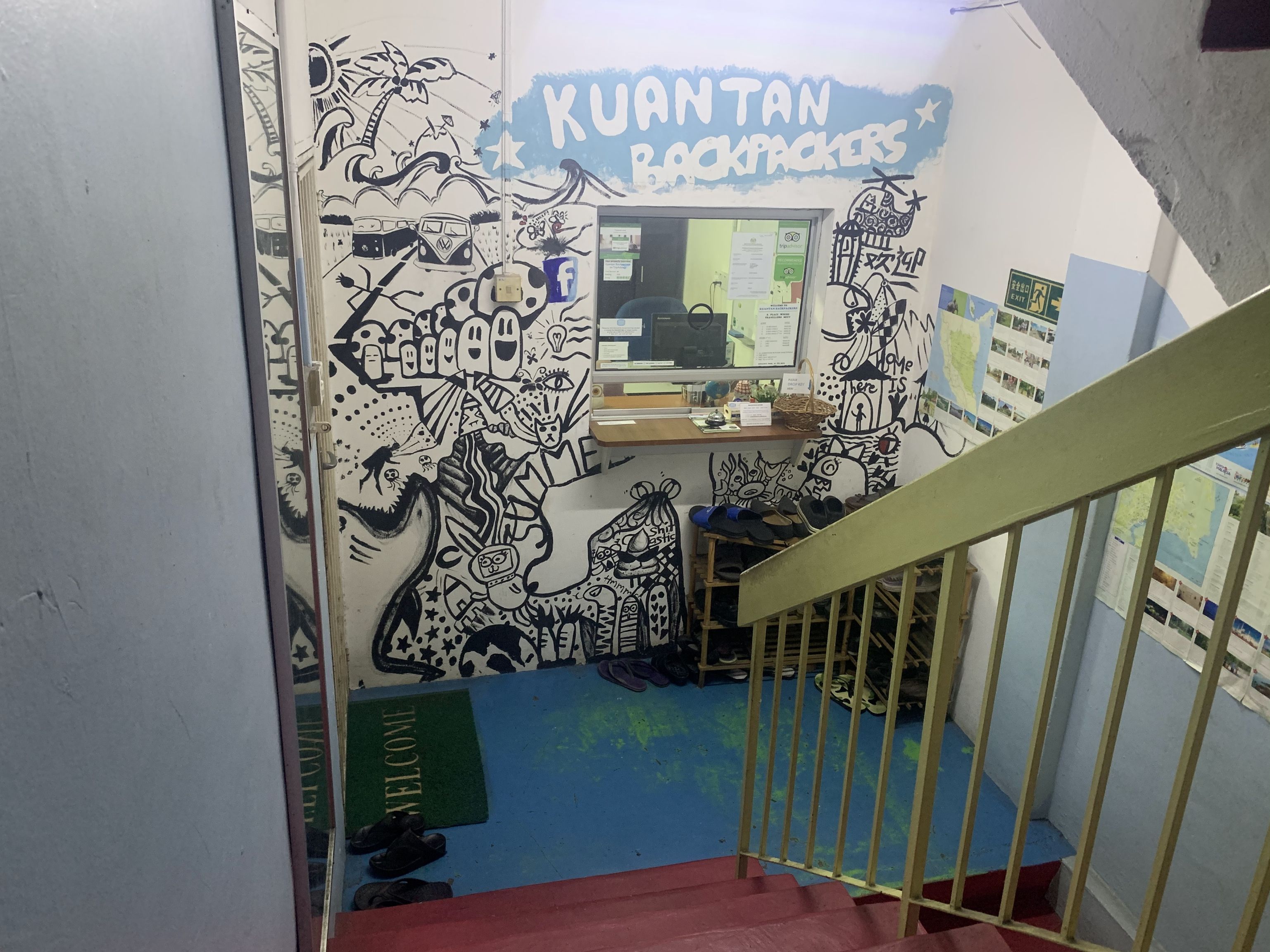 A staircase with a handrail, a wall with a mural titled 'KUANTAN BACKPACKERS,' and a desk with papers and a basket.