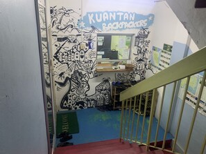 Una escalera con barandilla, una pared con un mural titulado 'KUANTAN BACKPACKERS' y un escritorio con papeles y una cesta.