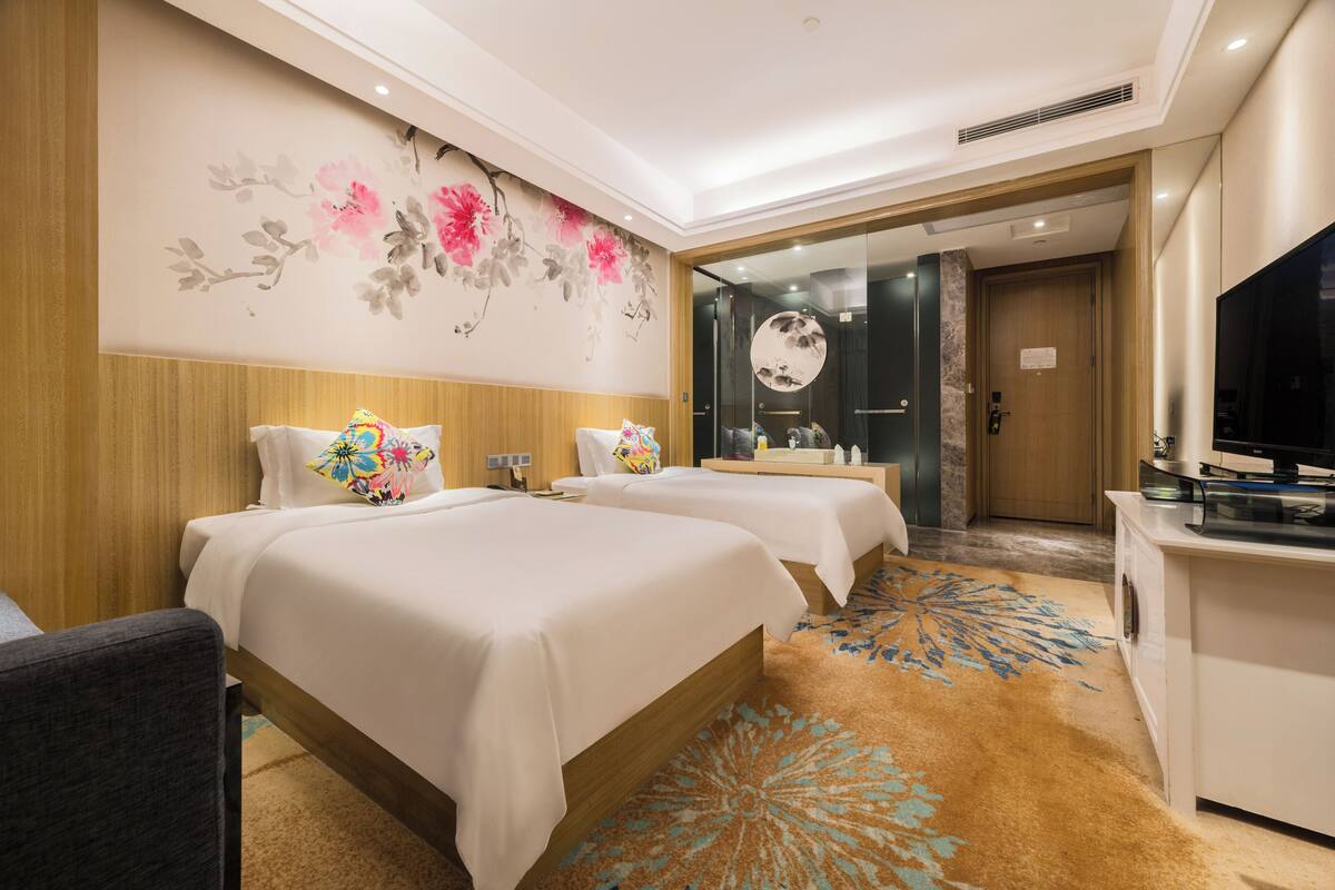 A hotel room with two beds, a desk, a TV, and a bathroom visible through a glass door.