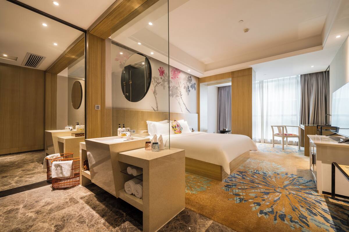 A hotel room with a bed, a desk, a chair, a bathroom with a mirror, and a large window.