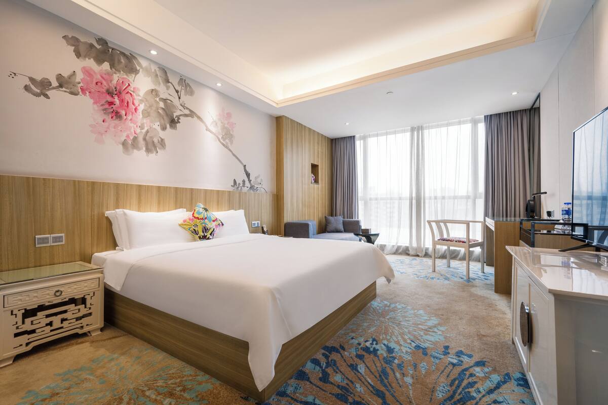 A hotel room with a large bed, a desk, a chair, a television, and a decorative wall painting.