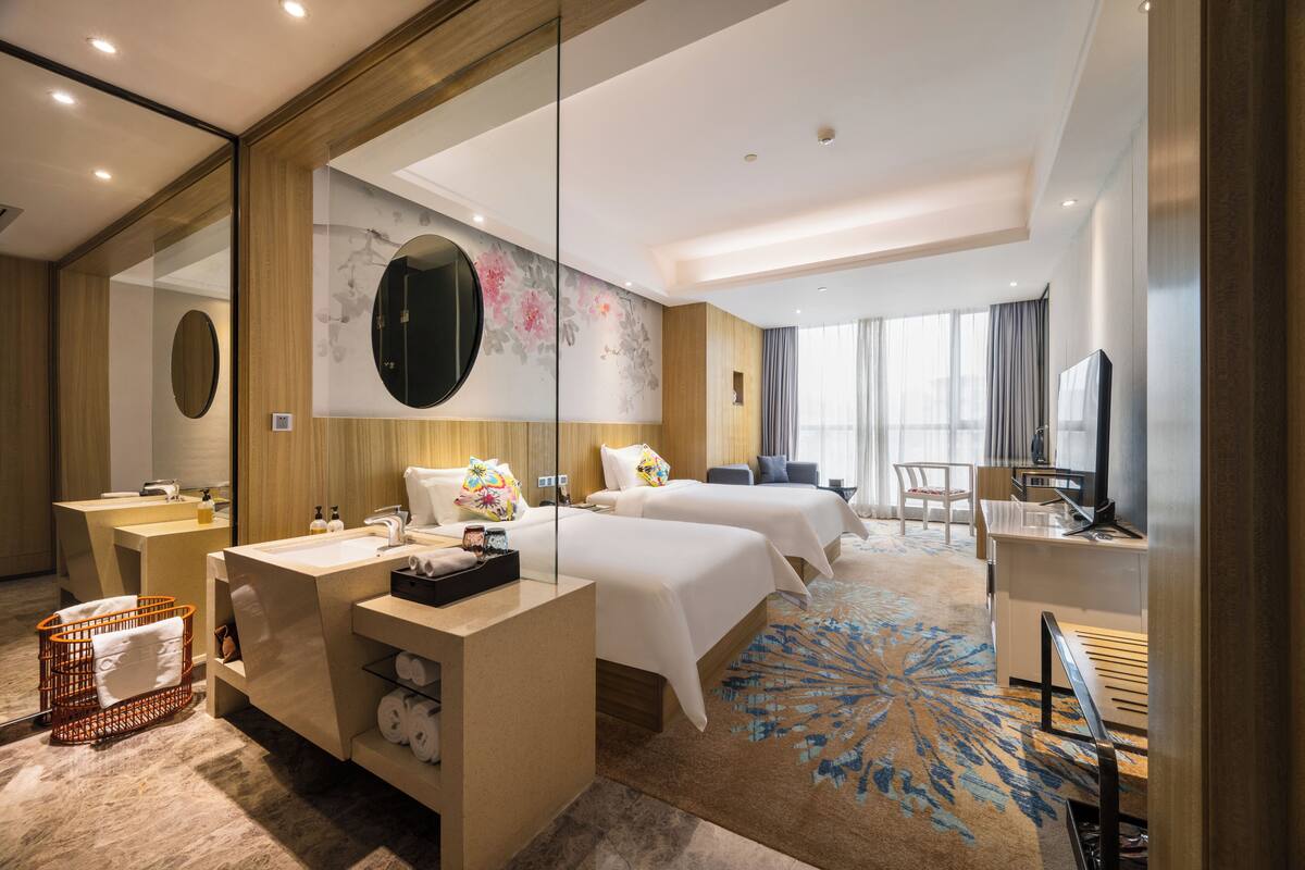 A hotel room with two beds, a desk, a TV, a large mirror, and a bathroom with a sink and toilet.
