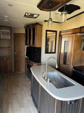 Una cocina con encimera de granito, pileta de acero inoxidable, muebles de madera, un microondas y un ventilador de techo.