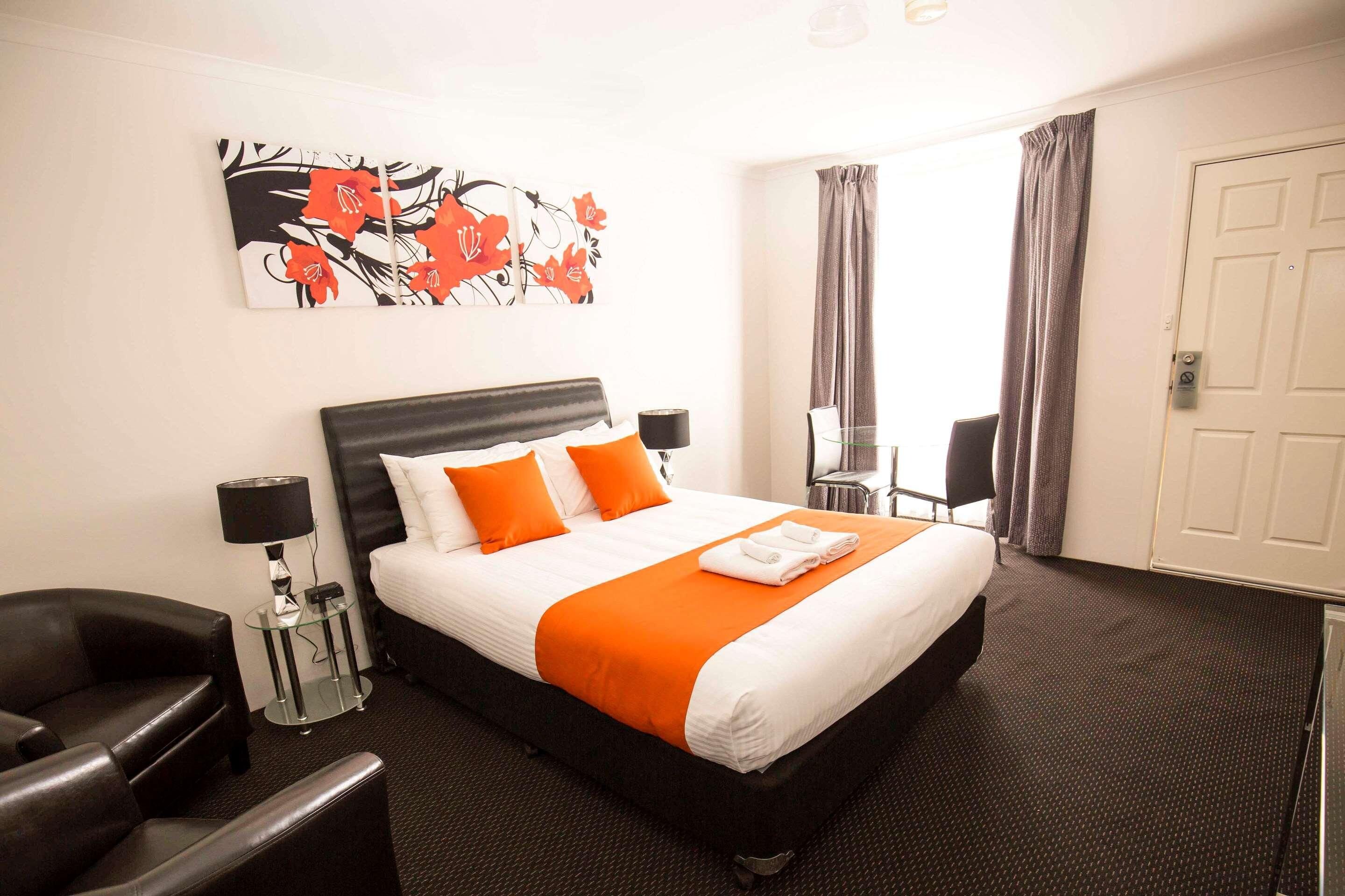 A hotel room with a bed, a chair, a small table with a lamp, a painting on the wall, and a door.
